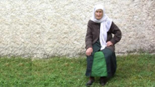 Eine ältere Frau mit Kopftuch sitzt auf einem Rasen vor einer Wand.