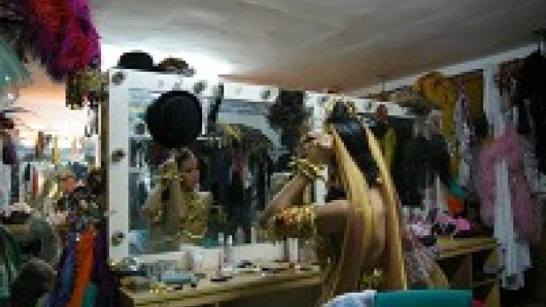 Eine Dragqueen schminkt sich vor einem Spiegel, umgeben von Kostümen und Accessoires.