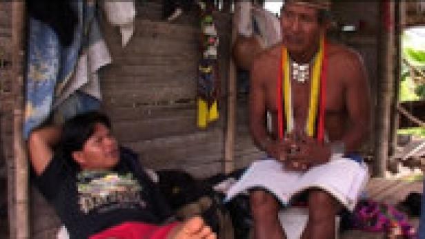 Zwei indigene Männer sitzen in einer einfachen Hütte, einer mit traditionellem Schmuck.