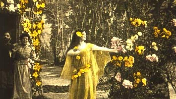 Eine Frau in einem gelben Kleid tanzt vor einem mit Blumen geschmückten Hintergrund.