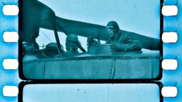 Ein Pilot und zwei weitere Personen sitzen in einem alten Doppeldecker-Flugzeug.