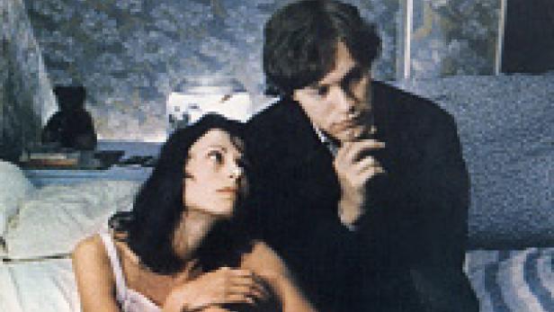 Eine Frau im Unterhemd und ein Mann im Anzug sitzen auf einem Bett.