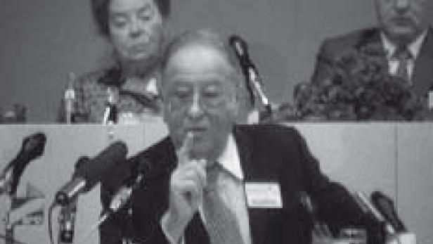 Ein Mann hält eine Rede vor Mikrofonen, während zwei Personen im Hintergrund sitzen.