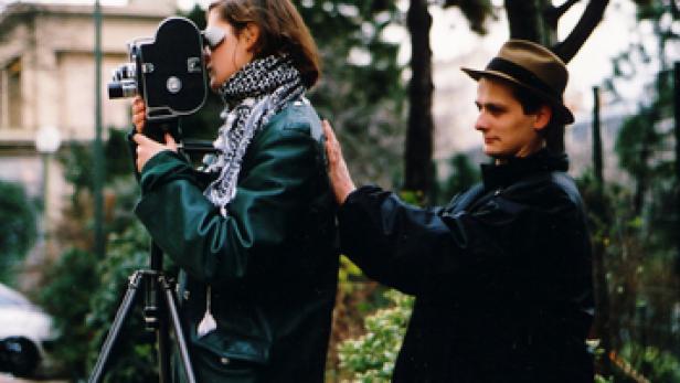 Eine Frau filmt mit einer alten Filmkamera, während ein Mann ihr die Hand auf den Rücken legt.