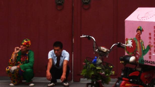 Ein Clown und ein Mann hocken vor einem roten Tor neben einem geschmückten Fahrrad.