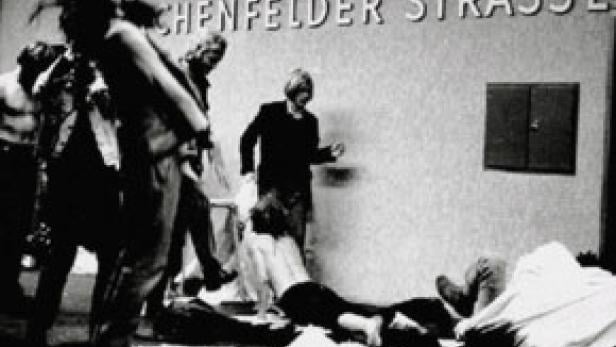 Eine Schwarzweißaufnahme von Menschen, die vor einer Wand mit der Aufschrift „Münchenfelder Strasse“ stehen.