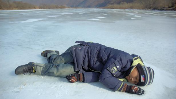 Ein Junge liegt auf einem zugefrorenen See, im Hintergrund sind Berge zu sehen.