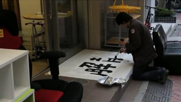 Ein Mann kniet und malt chinesische Schriftzeichen auf ein großes Blatt Papier.