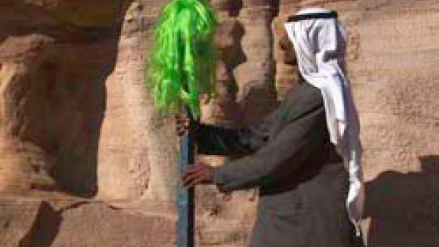 Ein Mann in traditioneller arabischer Kleidung hält eine grüne Perücke vor einer Felswand.