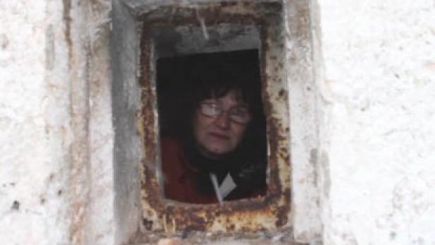 Eine Frau mit Brille schaut aus einer kleinen, rechteckigen Öffnung in einer Mauer.