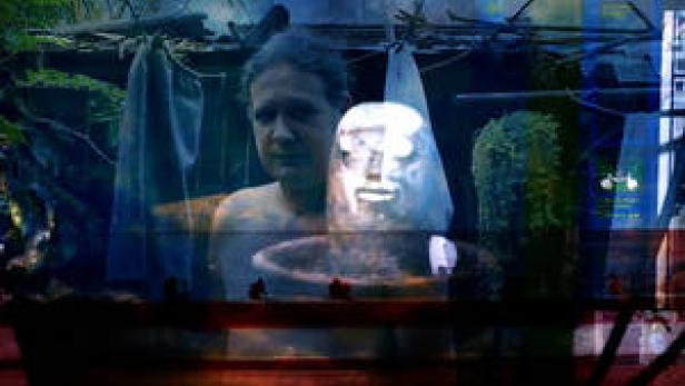 Ein Mann steht hinter einer silbernen Maske in einer dunklen Umgebung.