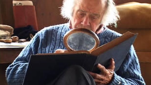 Ein älterer Mann mit weißem Haar betrachtet ein Buch durch eine Lupe.