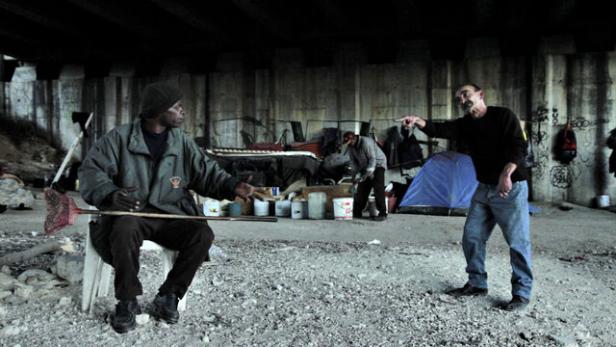 Unter einer Brücke sitzen und stehen mehrere Menschen in einem improvisierten Lager.