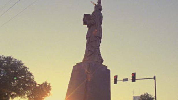 Eine Statue, die an die Freiheitsstatue erinnert, steht bei Sonnenuntergang in einer Stadt.