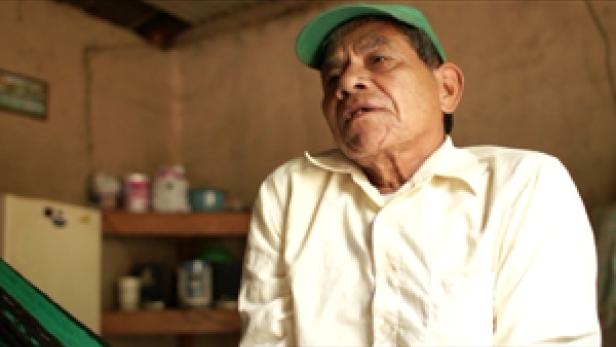 Ein älterer Mann mit grüner Kappe spricht in einem einfachen Raum.