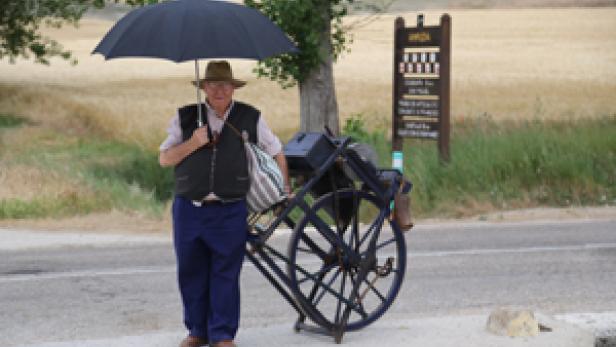 Ein älterer Mann mit Hut und Regenschirm steht neben einem beladenen Karren am Straßenrand.