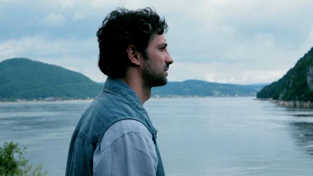 Ein Mann blickt auf einen ruhigen See, umgeben von bewaldeten Hügeln.