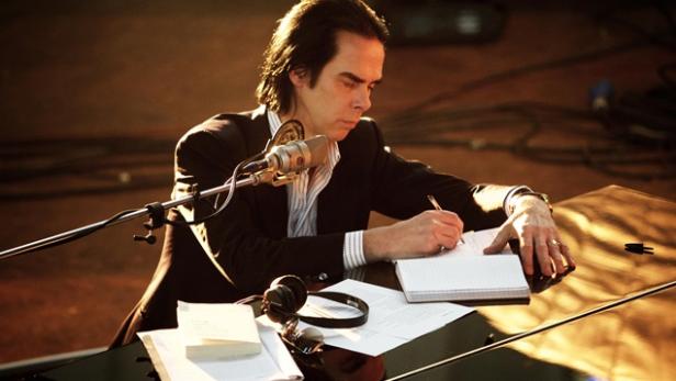 Nick Cave sitzt am Klavier und schreibt in ein Notizbuch.