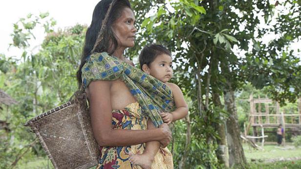 Eine Frau trägt ihr Kind in einer Tragevorrichtung durch eine ländliche Gegend.