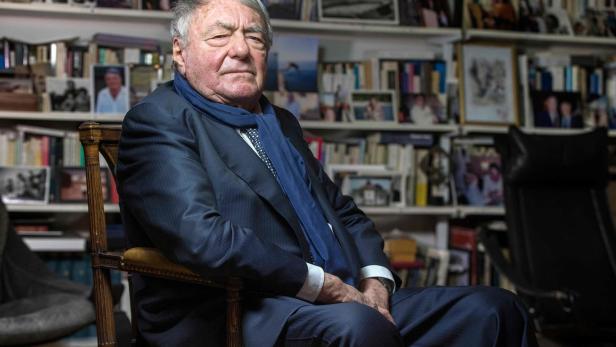 Ein älterer Mann mit Anzug und blauem Schal sitzt in einem Sessel vor einem Bücherregal.