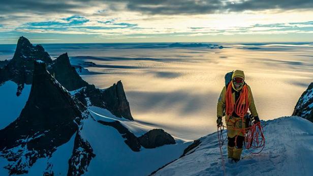 Ein Bergsteiger steht auf einem schneebedeckten Gipfel vor einer Berglandschaft.