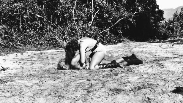 Zwei Frauen kämpfen auf einem sandigen Untergrund vor Büschen.