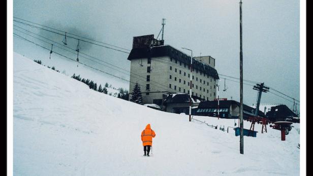 Ein Mann in oranger Jacke geht auf ein Hotel in einem verschneiten Skiort zu.