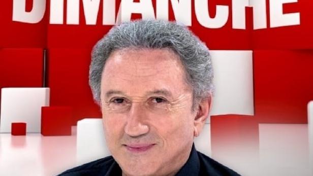 Ein Porträt von Michel Drucker vor dem Hintergrund der Sendung „Vivement Dimanche“.
