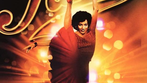 Eine Tänzerin in einem roten Kleid posiert vor dem Schriftzug „Fame“.