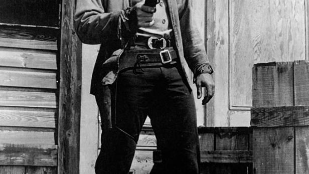 Ein Mann im Cowboyhut und mit Revolver vor einem Holzgebäude, im Vordergrund der Schriftzug „Bronco“.