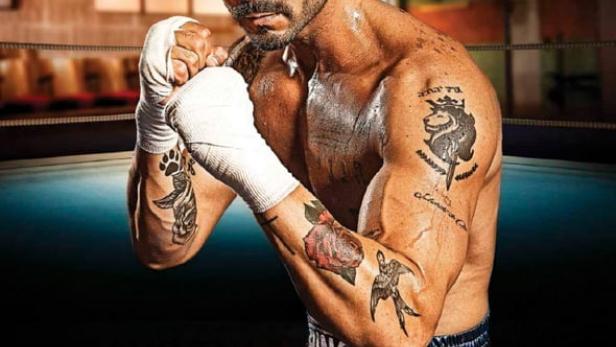 Ein muskulöser Boxer mit Tattoos und bandagierten Händen steht im Ring.