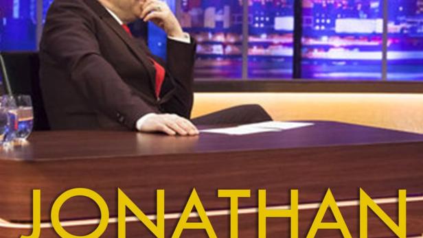 Jonathan Ross sitzt in seinem Studio vor einer beleuchteten Stadtkulisse.
