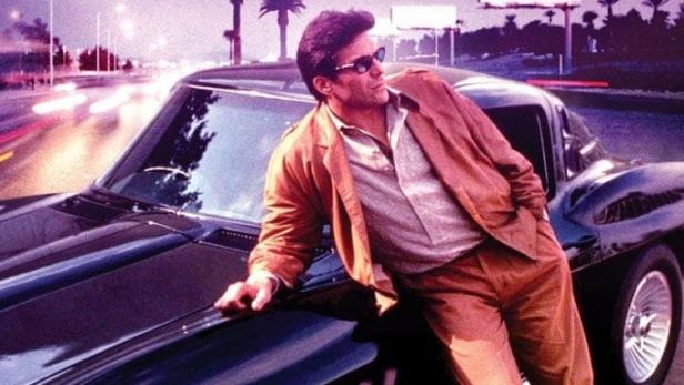 Ein Mann lehnt an einem schwarzen Corvette Stingray vor einer nächtlichen Stadtkulisse.