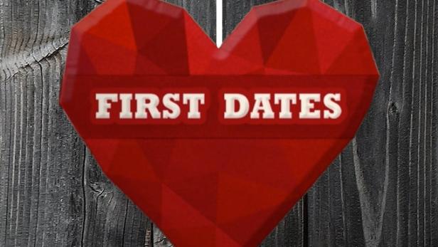 Ein rotes Herz mit der Aufschrift „First Dates“ hängt vor einer dunklen Holzwand.