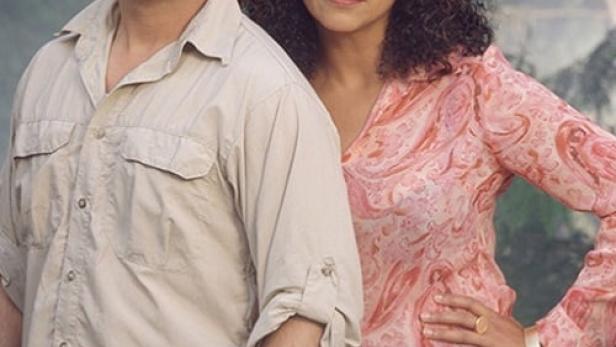 Ein Mann mit Brille und eine Frau mit lockigem Haar stehen vor einem grünen Hintergrund.