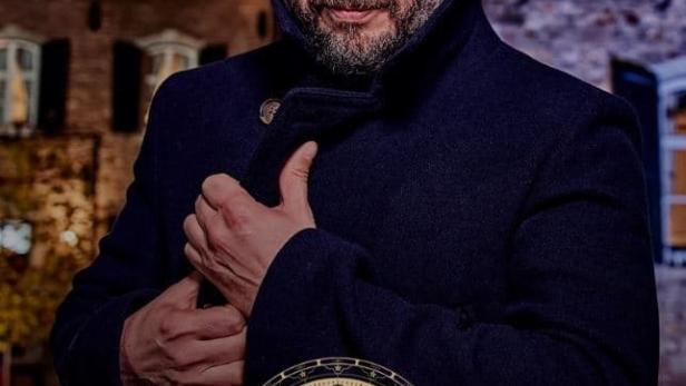 Ein Mann in einem dunklen Mantel vor einem Backsteingebäude, mit dem Logo von „De Verraders“.