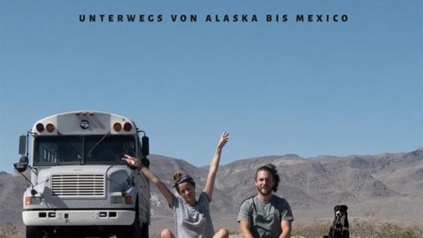 Ein Paar sitzt mit einem Hund und einem Bus auf einer Straße vor einer Bergkulisse; Titelbild von „Expedition Happiness“.