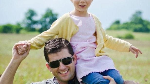 Ein Vater trägt seine kleine Tochter auf den Schultern durch eine Wiese.