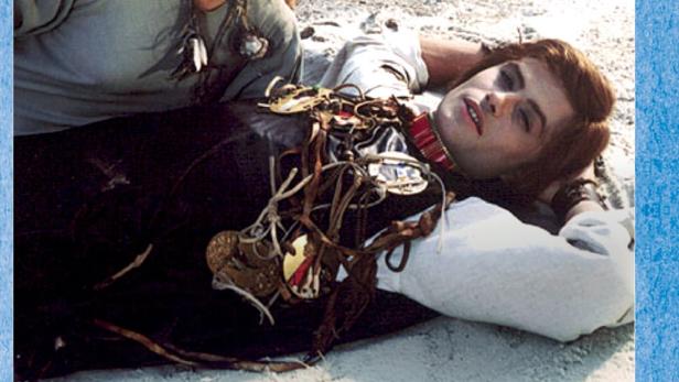 Eine Frau beugt sich über einen Mann, der mit Schmuck behangen am Strand liegt.