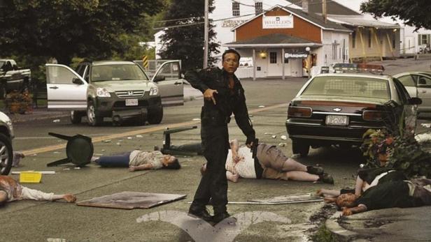 Eine Frau steht mit einer Waffe auf einer Straße voller Leichen, im Hintergrund ein Gebäude.