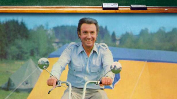 Ein Mann sitzt auf einem Motorrad vor einem Zeltplatz, über ihm steht „Hauptsache Ferien“ auf einer Tafel.