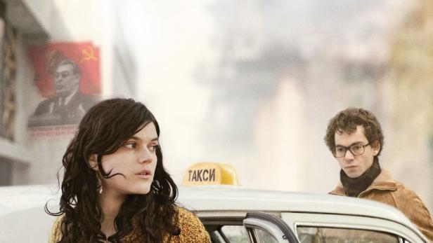 Eine Frau steigt aus einem Taxi vor dem Hintergrund eines sowjetischen Propaganda-Posters.