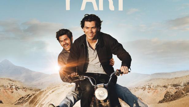 Zwei Männer fahren auf einem Motorrad durch eine wüstenähnliche Landschaft.