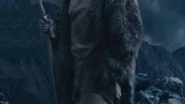 Ein Mann im Fellmantel hält einen Speer in der Hand, im Hintergrund eine Berglandschaft.