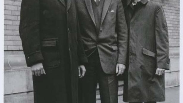 Drei Männer in Hüten und Mänteln stehen vor einem Gebäude der Victoria Police.