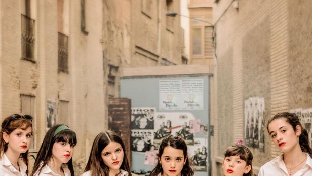 Sechs Mädchen in Schuluniformen stehen in einer Gasse vor Plakaten.