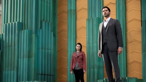 Tom Ellis als Lucifer Morningstar mit einer Frau im Hintergrund vor einem Gebäude.
