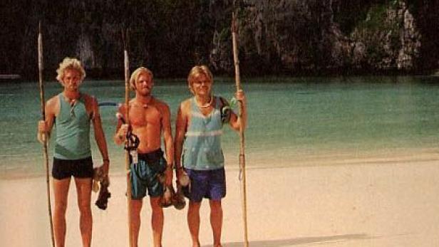 Drei Männer stehen mit Speeren am Strand vor einer Felswand und dem Meer.