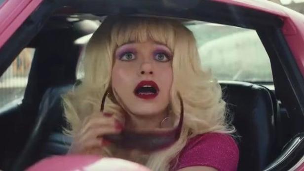 Eine Frau mit blonder Perücke sitzt überrascht in einem pinkfarbenen Auto.