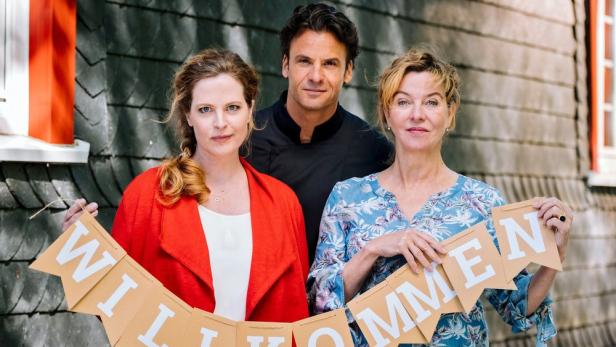 Drei Personen halten eine Girlande mit der Aufschrift „Willkommen“ vor einem Haus.